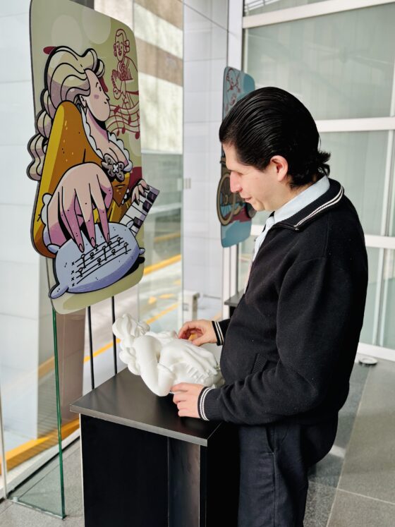 Foto vertical en perspectiva de la exposición "los muros que derribamos" donde aparece un hombre con discapacidad visual tocando la réplica táctil exhibida de la pianista ciega Maria Theresia Von Paradis.