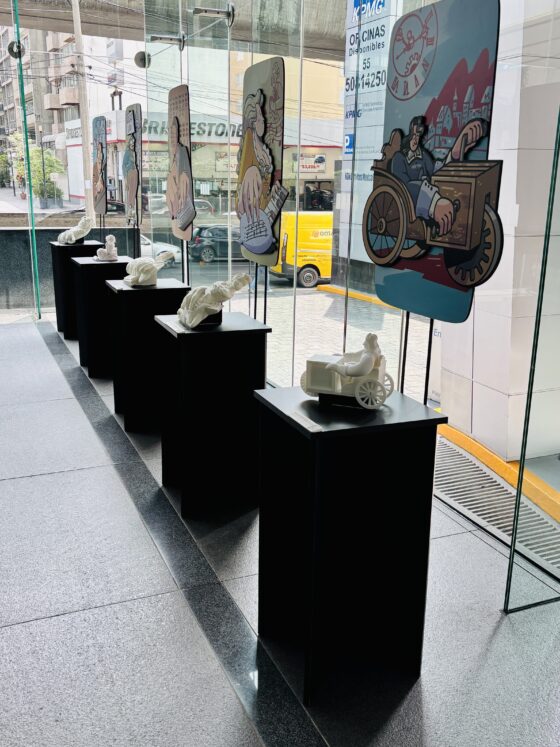 Foto vertical en perspectiva de la exposición "los muros que derribamos" instalada en el lobby de un corporativo. Se observan 5 módulos rectangulares de color negro, sobre los que se exhiben dibujos de personajes y sus respectivas réplicas táctiles impresas en 3D.