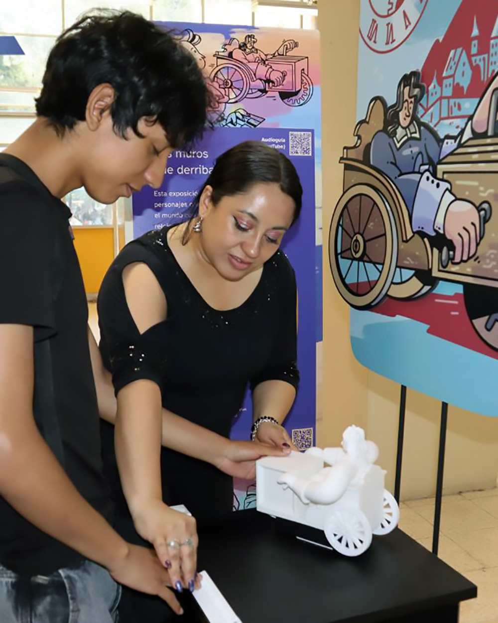 Foto vertical de dos personas, una maestra y un alumno, explorando con sus manos una figura impresa en 3D que representa a Stephan Farffler. Al fondo se ve una ilustración del mismo personaje.