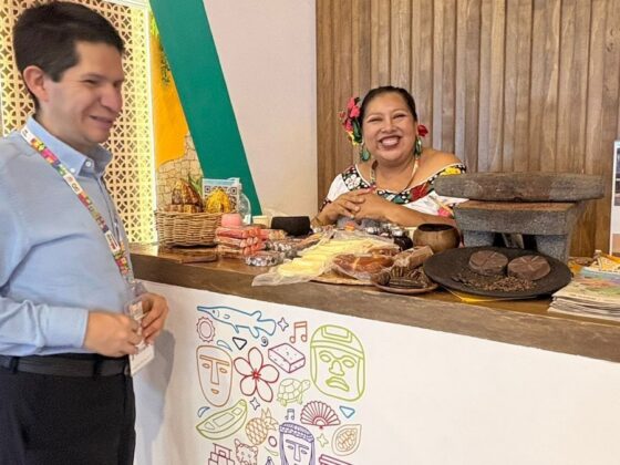 Foto vertical donde se observa a Cristóbal Sánchez, director del imetac, parado enfrente de un mostrador del tianguis, el cual tiene distintos productos de chocolate en exhibición arriba del mostrador en el cual lo está atendiendo una señora sonriente con la vestimenta típica tabasqueña.