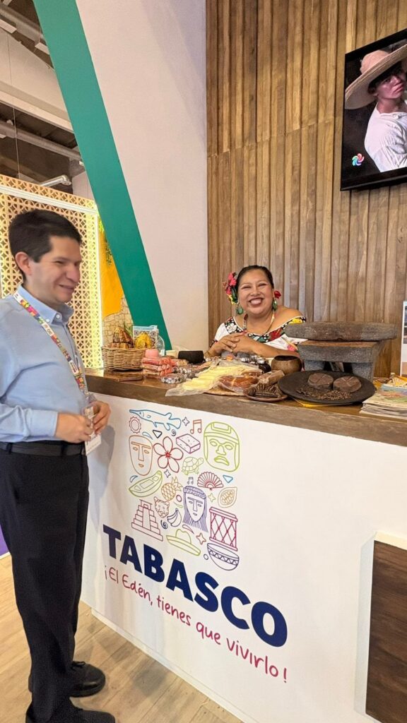 Foto vertical donde se observa a Cristóbal Sánchez, director del imetac, parado enfrente de un mostrador del tianguis, el cual tiene distintos productos de chocolate en exhibición arriba del mostrador en el cual lo está atendiendo una señora sonriente con la vestimenta típica tabasqueña.