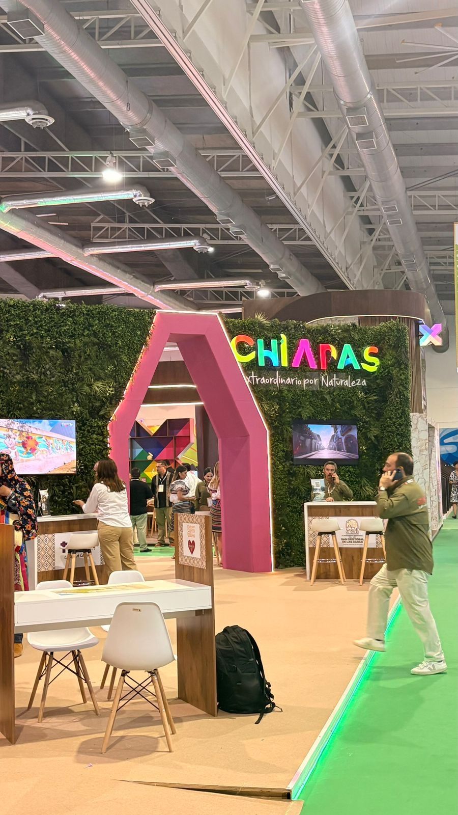 Foto vertical de una parte del tianguis donde se observa un puesto relacionado con Chiapas el cual lo atiende un señor que está sentado hablando por teléfono, se observa un señor caminando por el lugar y otras dos personas en otro puesto.