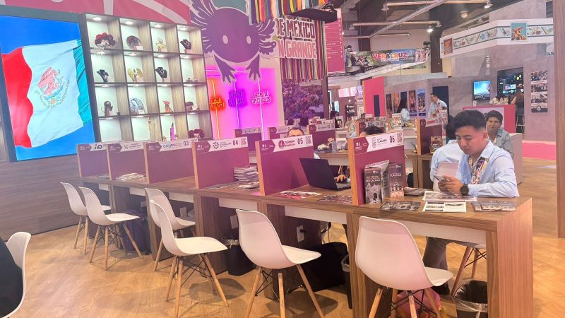 Fotografía vertical que muestra un módulo de atención dentro del tianguis turístico. La estructura del stand es de madera clara y cuenta con una barra larga dividida en secciones numeradas mediante carteles de color guinda, cada una equipada con una silla blanca. En el extremo derecho de la barra, una persona con camisa clara revisa unos folletos informativos. De fondo una pantalla que proyecta la bandera de México, junto a un estante con diferentes piezas artesanales y a un costado un logotipo con la silueta de un ajolote.