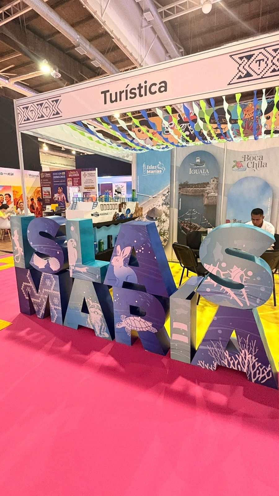 Fotografía vertical que muestra un stand promocional en el tianguis turístico con el letrero superior "Turística". En el centro de la escena, destacan letras tridimensionales de gran tamaño que forman la frase "ISLAS MARÍAS". Las letras están decoradas con ilustraciones de la fauna marina incluyendo una tortuga y un tiburón sobre un fondo azul que simula el océano. Al fondo, el stand cuenta con mamparas informativas que muestran imágenes de paisajes costeros, logotipos de destinos como "Islas Marías", "Iguala" y "Boca de Chila" y se observa a un expositor sentado atendiendo el espacio.