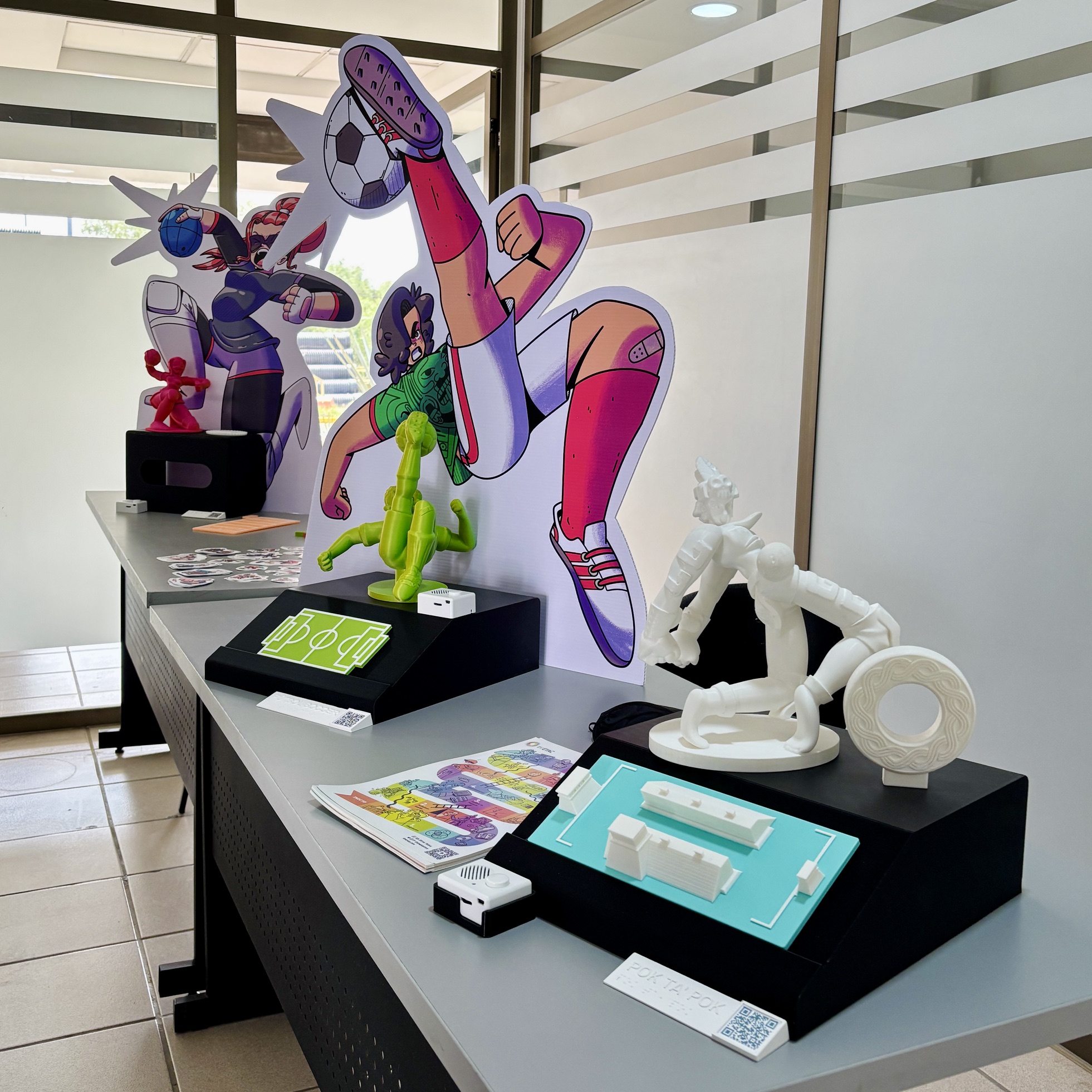 Foto de la exposición accesible “de bote pronto” del imetac montada en una oficina durante la semana nacional de la cultura del agua. Se conforma por piezas impresas en 3D de jugadores de juego de pelota prehispánico, fútbol soccer y golbol, sobre bases en las que también hay representaciones de las canchas, porterías, balones, cédulas en braille y bocinas con audio guías. Detrás de cada una hay ilustraciones grandes de los personajes.