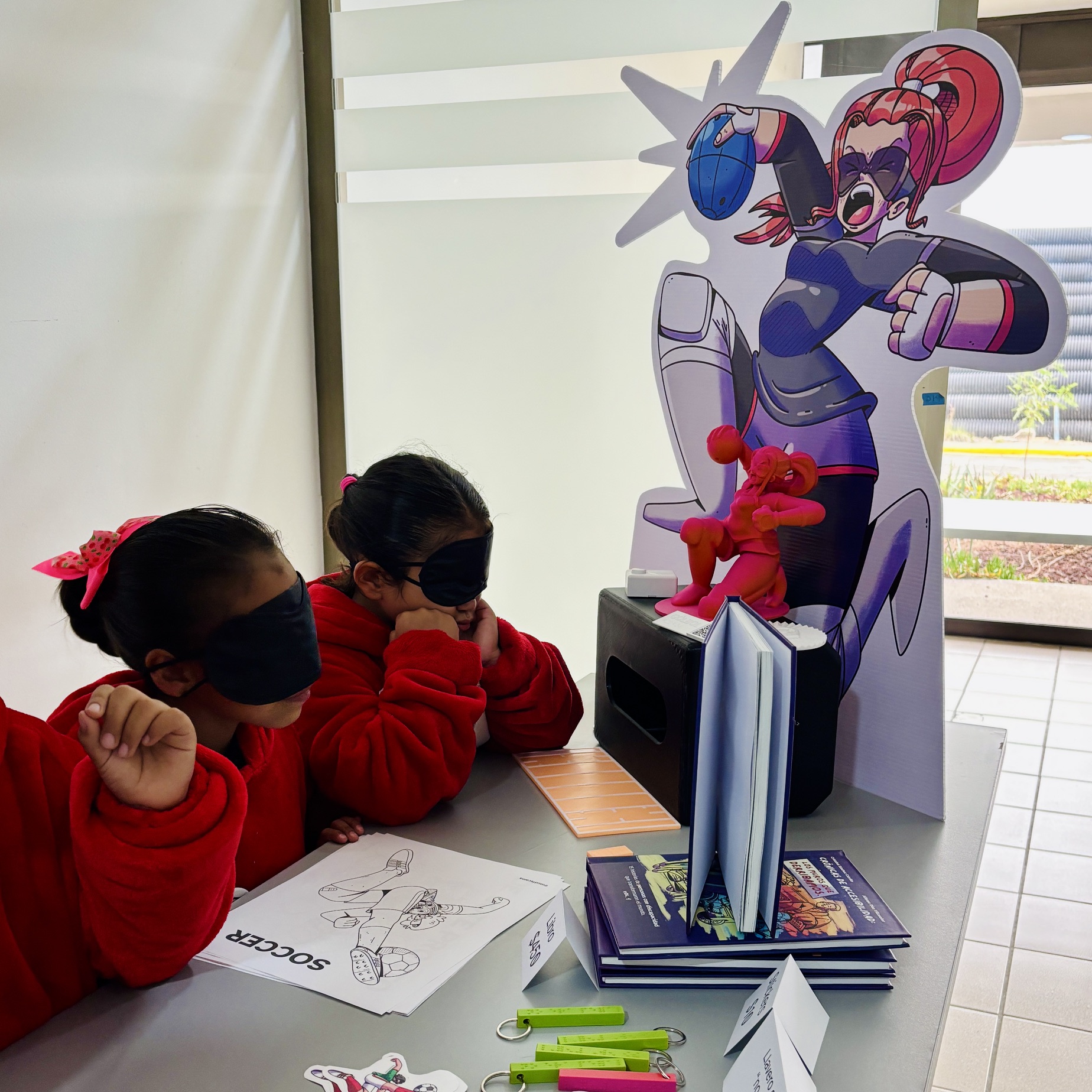 Foto de dos niñas con antifaces escuchando una audioguía sobre un deporte accesible llamado gólbol. Frente a ellas hay una jugadora de este deporte impresa 3D, su respectiva ilustración en gran formato, y a su lado una réplica de una medalla paralímpica.