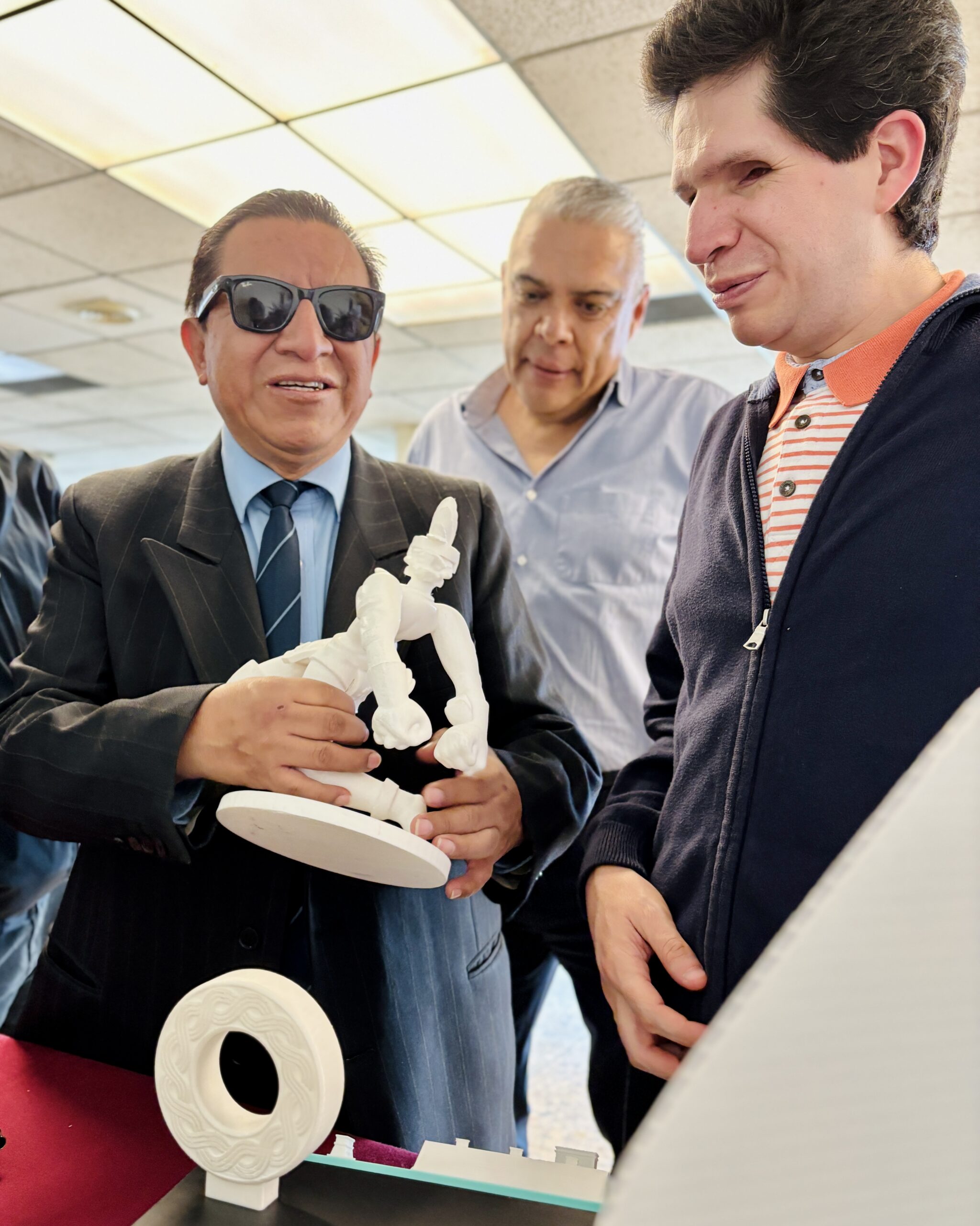 Foto vertical de Cristóbal Sánchez, director general del IMETAC, mostrándole a otra persona con discapacidad visual que se encuentra a su derecha una impresión 3d de un jugador de juego de pelota prehispánico. La persona con discapacidad visual que se encuentra con él es un hombre que viste traje formal, corbata y gafas de sol oscuras mientras sostiene con ambas manos la impresión 3d de color blanca. Entre medio de ellos dos un poco atrás otro hombre con camisa clara observando lo que ellos hacen.