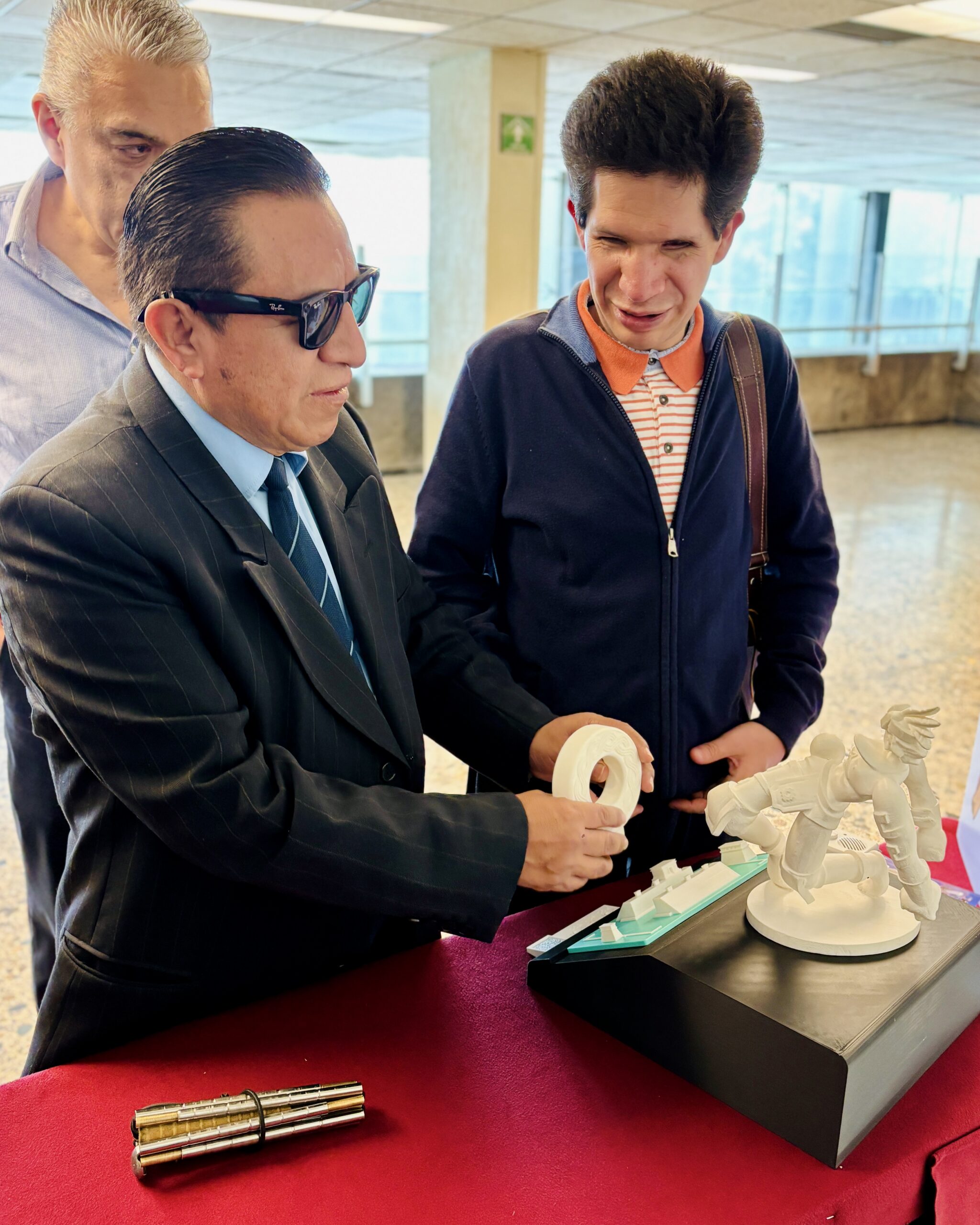 Foto de dos personas con discapacidad visual tocando impresiones 3d de la cancha y aros del juego de pelota prehispánico. La primera persona se encuentra a la izquierda, un hombre con traje oscuro, corbata azul y lentes de sol sosteniendo una impresión 3d, A un lado se encuentra de Cristóbal Sánchez, director general del IMETAC, con una chaqueta azul y camisa de rayas. Frente a ellos una mesa con un mantel rojo y encima de la mesa se encuentran las impresiones 3d que están tocando.