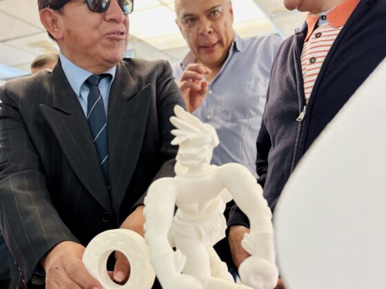 Foto de Cristóbal Sánchez mostrando la exposición accesible “de bote pronto” a 2 personas. Frente a ellos aparecen varias impresiones 3d de color blanco en una mesa las cuales están examinando. El hombre de la izquierda viste traje formal, corbata y gafas de sol oscuras mientras toca una pequeña pieza circular, Cristóbal viste con una chaqueta azul y camisa de rayas, la tercera persona entre medio de ellos dos viste camisa clara En el centro de la mesa resalta una figura humana de color blanca.