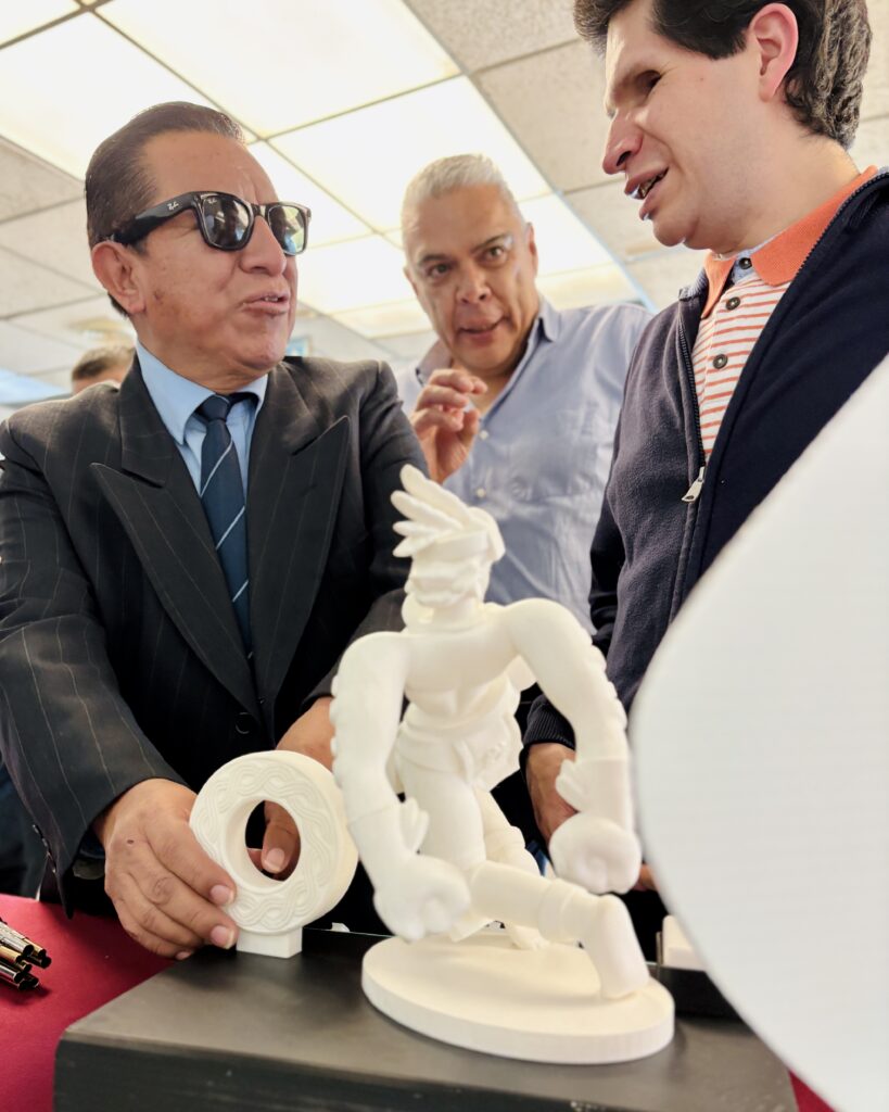 Foto de Cristóbal Sánchez mostrando la exposición accesible “de bote pronto” a 2 personas. Frente a ellos aparecen varias impresiones 3d de color blanco en una mesa las cuales están examinando. El hombre de la izquierda viste traje formal, corbata y gafas de sol oscuras mientras toca una pequeña pieza circular, Cristóbal viste con una chaqueta azul y camisa de rayas, la tercera persona entre medio de ellos dos viste camisa clara En el centro de la mesa resalta una figura humana de color blanca.