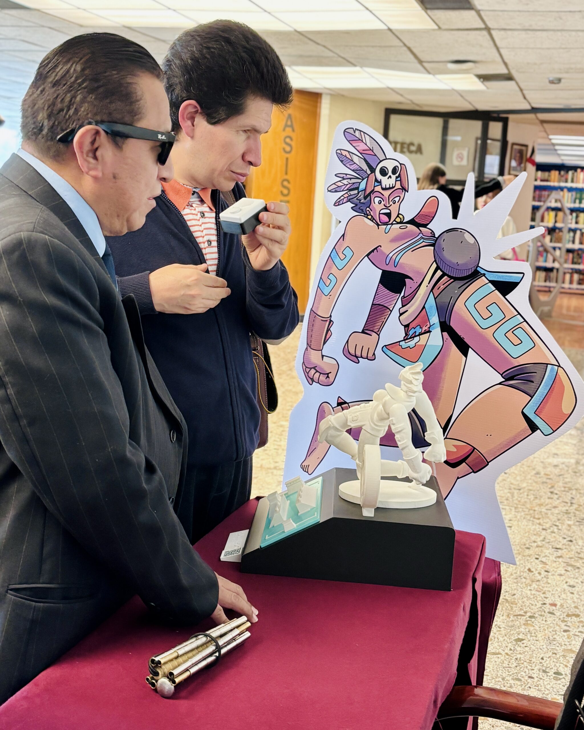 Foto de dos personas con discapacidad visual escuchando una audio guía sobre el juego de pelota prehispánico. A su lado aparece una ilustración grande de un jugador y su respectiva réplica impresa en 3d. uno de ellos viste un traje oscuro y gafas, mientras que el otro usa una chaqueta azul y sostiene la audio guía cerca de su rostro.