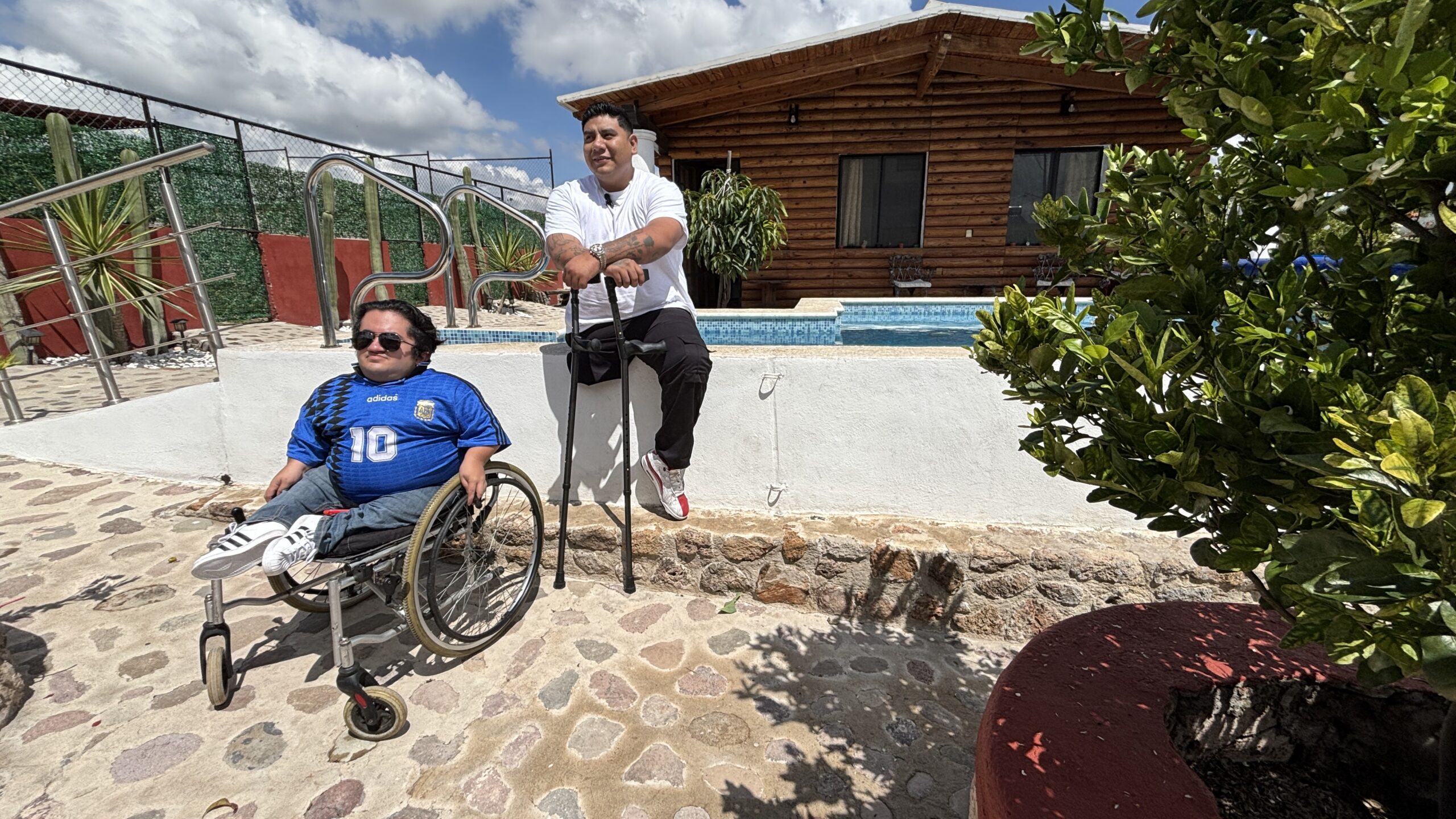 Imagen horizontal de la fachada de Villa Bugambilia, una cabaña con medidas de accesibilidad implementadas de manera integral. Al frente aparecen dos viajeros con discapacidad motriz, uno usuario de silla de ruedas y otro con amputación en una de sus piernas.